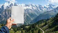 Caucasian male hand holding blank white card against alpine mountain landscape with snow peaks, forested valleys, and winding roads for mockup and advertising purposes.