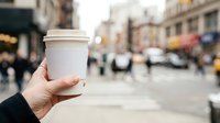 Hand holding white takeaway coffee cup on urban street with blurred city background and pedestrians, suitable for beverage, lifestyle, and commercial use.