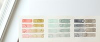 Watercolor paint swatches in soft pastel colors on white paper showing various brush strokes and color mixing techniques for artistic projects.