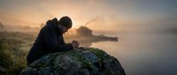 Contemplative man sitting on mossy rock by misty lake at sunrise, peaceful morning meditation scene with soft golden light and serene atmosphere.