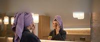 Young woman with purple hair towel applying skincare routine in modern bathroom mirror reflection for wellness and self-care morning ritual.