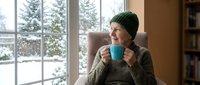Young woman in green knit beanie and sweater enjoying hot drink by window with snowy winter landscape view. Cozy indoor lifestyle moment for seasonal content.