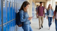 Teenage girl using smartphone by school lockers while students walk in hallway background during break time.