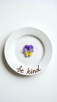 White ceramic plate with a purple yellow pansy flower and be kind handwritten text, minimalist inspirational concept on white background.
