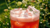 Refreshing iced pink drink in a glass with ice cubes, set against a lush green garden background on a sunny summer day.