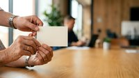 Man holding blank business card at wooden table in modern office environment, suitable for corporate branding, networking, and professional identity design projects.