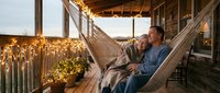 Mature man and woman relaxing together in hammock on rustic wooden porch with warm string lights and greenery, enjoying peaceful evening outdoors for lifestyle and relaxation concepts.