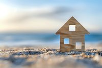 Wooden house miniature on sandy beach against blurred ocean background, symbolizing coastal living, vacation homes and real estate investment dreams.