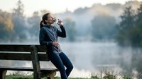 Young Asian woman drinking coffee while sitting on wooden bench by misty lake during peaceful morning outdoors in autumn park setting.