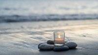 Peaceful beach scene with glowing candle in glass holder surrounded by smooth dark stones on wet sand with soft ocean waves and cloudy sky at sunset.