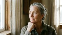 Mature woman with gray hair and fair complexion, wearing olive-toned cardigan, resting chin on hand in contemplative pose near window with natural light and neutral curtains.