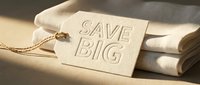 Beige clothing tag with Save Big text resting on folded fabric in warm sunlight, promoting retail sale and discount shopping concepts.