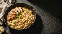 Delicious spaghetti pasta with grilled chicken breast and fresh herbs served in cast iron skillet on dark stone surface for culinary presentation.
