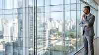 Mature male professional in gray business suit standing by floor-to-ceiling windows in modern high-rise office, contemplating cityscape view, suitable for corporate leadership and business concepts.