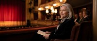 Elegant senior woman with blonde hair reading program in ornate theater with red curtains and golden lighting during cultural performance.
