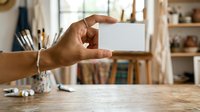 Person with medium skin tone holding blank white card at wooden desk with office supplies and bookshelves, suitable for business branding, corporate identity, and marketing materials.