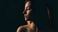 Young woman in dramatic side profile with warm golden lighting against dark background creating moody atmospheric portrait for beauty and wellness concepts.