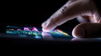 Iridescent crystal formation on dark surface with human finger touching delicate rainbow-colored mineral structures in dramatic lighting and macro photography.