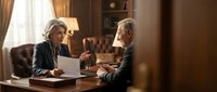 Two mature professionals in business attire discuss documents at wooden desk in warm office setting with lamp and shelving, suitable for corporate, legal, and business communication concepts.