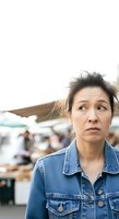Middle-aged East Asian woman wearing denim jacket looking confused or concerned at outdoor market or street fair with blurred crowd background.