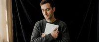 Young Caucasian man in gray knit sweater holding white tablet device while sitting indoors, casual lifestyle portrait for technology and digital communication concepts.