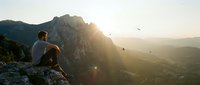 Hiker sitting on mountain cliff edge at sunrise, contemplating vast valley landscape with golden light and soaring birds below, adventure and freedom concept.