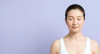Middle-aged Asian woman with dark hair pulled back, wearing white sleeveless top, eyes closed in peaceful expression against lavender background for wellness and mindfulness content.
