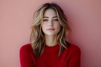 Young Caucasian woman with highlighted blonde hair wearing red knit sweater smiling confidently against pink background, natural makeup and modern casual style.