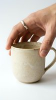 Hand with silver ring holding a rustic handmade ceramic mug with speckled glaze on white background, artisan pottery concept.