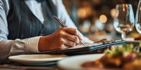 Female server taking order at upscale restaurant with clipboard and pen, elegant dining atmosphere with wine glasses and gourmet food.