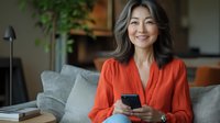 Mature Asian woman in bright orange blouse smiling while using smartphone on couch. Natural gray hair and casual home office setting with indoor plants and modern decor.