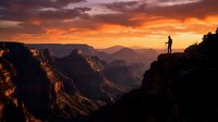 Silhouetted hiker stands on rocky cliff edge overlooking vast canyon landscape during dramatic golden sunset with layered mountain peaks and glowing orange sky.