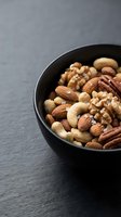 Mixed nuts assortment in black bowl on dark slate surface featuring almonds, walnuts, cashews and pecans for healthy snacking and cooking.