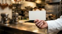 Person holding blank white card in restaurant kitchen environment with warm lighting and blurred background.