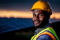 African American male worker in safety vest and hard hat smiling at solar panel installation site during sunset, representing renewable energy and sustainable development.