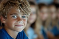 Young blonde boy with blue eyes smiling warmly in classroom setting with blurred students in background for education and childhood concepts.