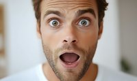 Young Caucasian man with beard expressing extreme surprise with wide open eyes and mouth, wearing white t-shirt against light background. Close-up portrait showing genuine emotion.