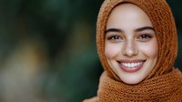 Young Middle Eastern woman wearing brown hijab smiling warmly at camera with natural makeup and white teeth against blurred green background.