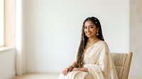 Young woman with long braided hair and medium brown skin tone wearing a cream-colored linen shirt, seated on a natural woven chair in a bright, minimalist interior space with white walls and natural light.