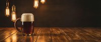Dark beer glass with foam on rustic wooden table under warm vintage Edison bulb lighting creating cozy pub atmosphere for brewery marketing materials.