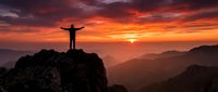 Man celebrating success on mountain peak at dramatic sunset with arms raised against orange sky and layered mountain ranges in distance.