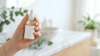 Unisex hand holding blank white cosmetic bottle in bright bathroom with green plants and natural light for skincare product display.