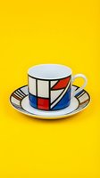 Porcelain teacup and saucer with geometric abstract design featuring red, blue, and black shapes on bright yellow background. Isolated tableware.