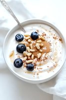 Greek yogurt bowl topped with fresh blueberries, pine nuts, almond butter and cinnamon powder on white ceramic plate, minimalist breakfast styling.