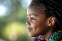 Young African girl with braided hair smiling in profile view against blurred natural background, wearing colorful traditional pattern clothing, expressing joy and innocence.