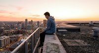 Young man sitting on rooftop edge overlooking city skyline at sunset, urban exploration and contemplation scene for lifestyle and travel content.