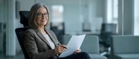 Mature woman with gray hair and glasses wearing brown blazer holding laptop in modern office environment with blurred background.