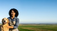 Man with curly dark hair and Middle Eastern ethnicity posing outdoors with golden retriever dog against expansive agricultural landscape and clear blue sky.