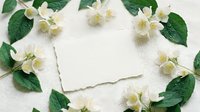 Blank white card surrounded by fresh jasmine flowers and green leaves on neutral linen fabric background, suitable for wedding invitations, greeting cards, and stationery design projects.