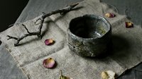 Rustic ceramic bowl with moss texture on burlap fabric surrounded by dried rose petals and twigs creating natural organic still life composition.